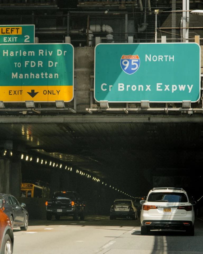 A view from a highway looking directly into a dark underpass or tunnel, with several large green overhead traffic signs mounted above the entrance. The left sign reads "LEFT EXIT 2 / Harlem Riv Dr TO FDR Dr / Manhattan" with a yellow bottom section stating "EXIT ONLY" next to a downward arrow. The larger right sign features the Interstate 95 shield and reads "NORTH / 95 / Cr Bronx Expwy." Several vehicles, including a white SUV on the right and a black pickup truck on the left, are driving into the illuminated tunnel.