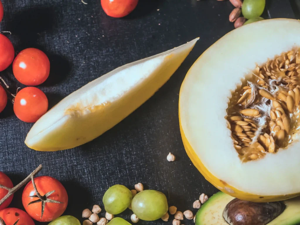 A vibrant, close-up photograph featuring an assortment of fresh produce that visually illustrates the rich aroma and tropical flavor profile of the CAM Devil Driver cannabis strain. In the center, a ripe yellow melon is cut open to reveal its seed-filled interior, accompanied by a single sliced wedge to represent the strain's prominent melon notes. Arranged across a dark, textured surface are plump green grapes, scattered chickpeas, a sliced avocado, and bright red cherry tomatoes. This fresh combination of sweet fruit elements beautifully mirrors the strain's complex bouquet of bold grape candy, sweet cream, and fruity cereal undertones