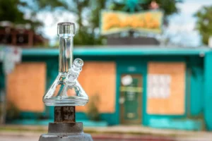 A crisp, eye-level product photograph featuring a small, clear glass beaker bong with the brand name 'GRAV' etched vertically down its neck. The pristine glass water pipe rests on top of a weathered, grey metallic post or fire hydrant in sharp focus in the foreground. In the softly blurred background sits a retro, single-story turquoise building featuring boarded-up wooden window panels and a yellow vintage sign under a bright sky. The sparkling clarity of the glass contrasts vividly against the colorful, urban backdrop.
