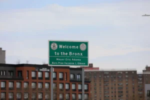A daytime, eye-level photograph of an official green municipal road sign welcoming visitors to New York City. The rectangular sign features bold white text that reads 'Welcome to the Bronx,' flanked by two circular official seals in the upper corners. Below a white dividing line, the sign lists the city's leadership, reading 'Mayor Eric Adams' and 'Boro Pres Vanessa L Gibson.' The sign is mounted on two perforated metal posts. In the background, multi-story brick apartment buildings and urban residential structures stand beneath a soft, overcast sky.