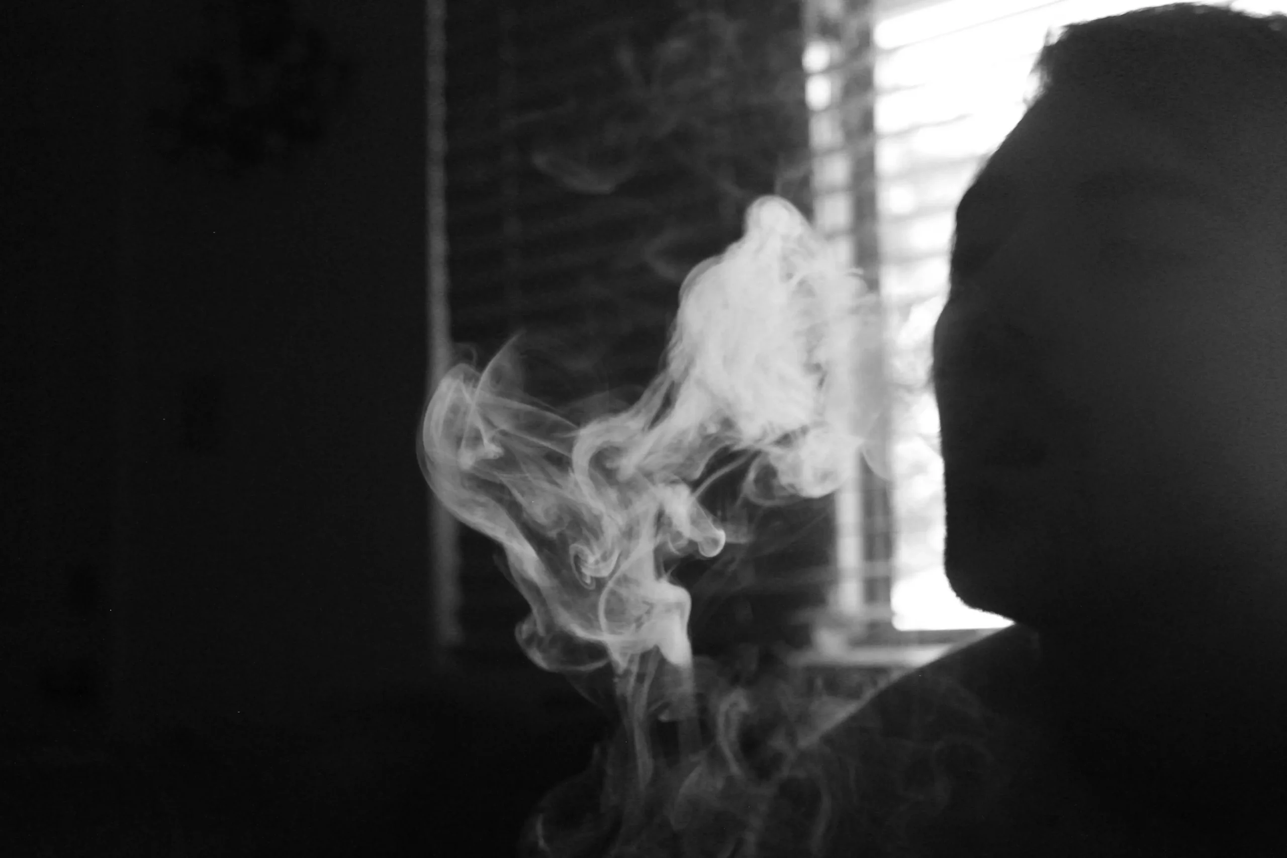 An atmospheric, sunlit photograph capturing a close-up of a person's hand holding a lit joint with a thin, delicate stream of smoke curling upward from its ash-covered tip. The hand is positioned over a wooden table spread with bohemian and creative essentials, including a dish holding a white sage smudge stick and Palo Santo wood. On the left, a lush, vibrant green fern catches bright natural light coming from a nearby window, while art supplies and a small crystal cluster are softly blurred in the background, creating a relaxed, creative studio environment.